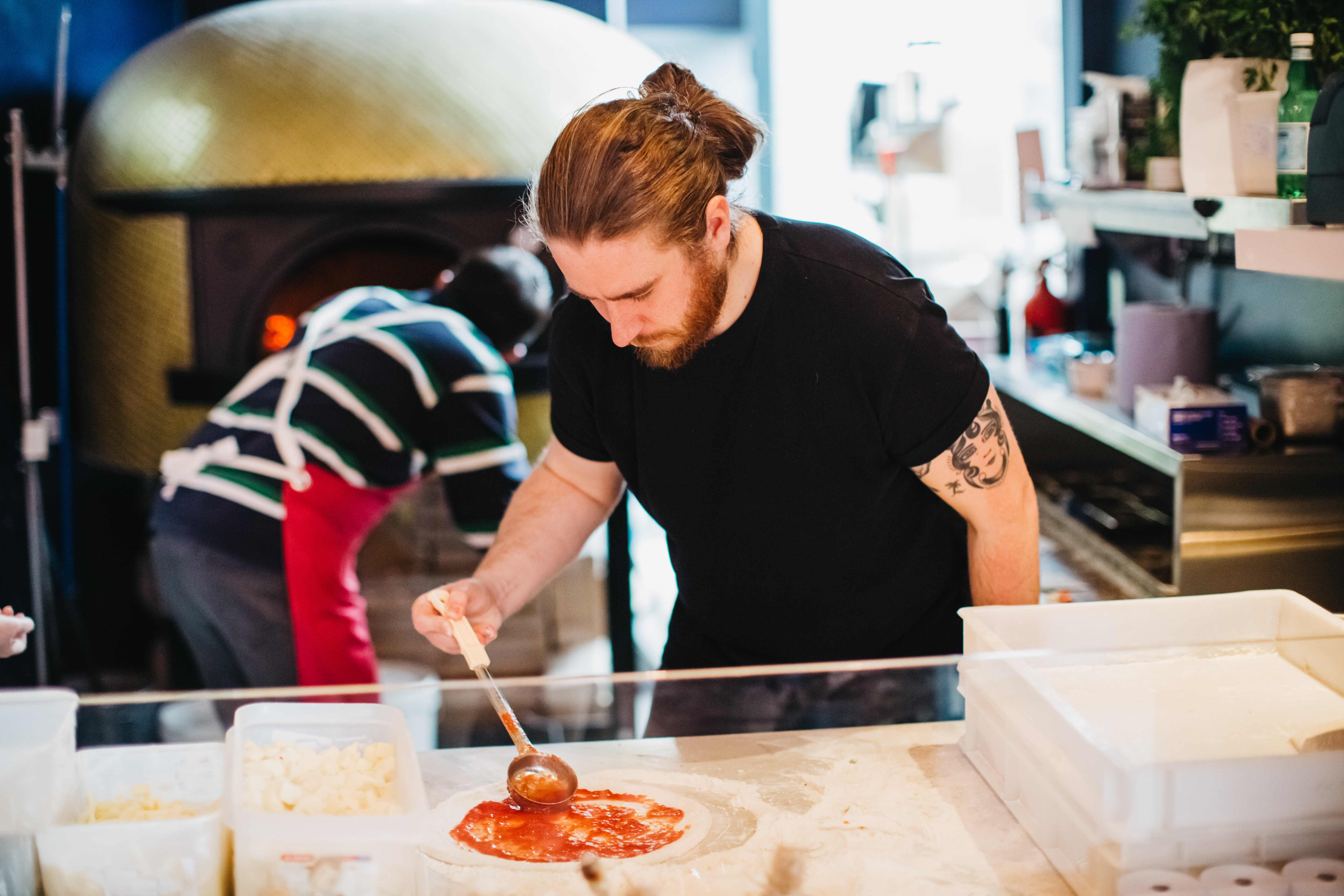 Man in a black t-shirt adding sauce to a pizza base